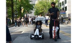 A man on a scooter carries a protest sign while a little girl rides a miniature car alongside him