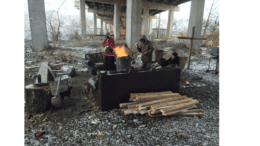 A homeless camp under an overpass with three men around a fire in a steel drum