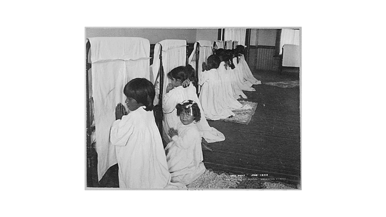 Unidentified Native American girls at the Phoenix Indian School in June 1900 pray beside their beds. Photo via National Archives
