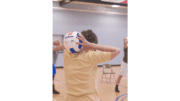 The older individuals pictured here, on the wooden floor of an indoor basketball court, were participating in an exercise class consisting of stretching, and aerobic repetitive motion movements. At this point in their exercise session, the participants were working the muscles of their arms and shoulders, by holding an inflatable volleyball with both hands, and lifting the ball over, and behind their head, resting it on their neck. This exercise also helped to stretch the muscles as well.