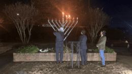 Two people stand to the side as a man lights the third candle of a Menorah