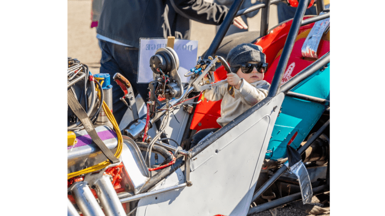 A small child sits in the open cab of a drag racer. The child is wearing a wool pullover cap and large sun glasses