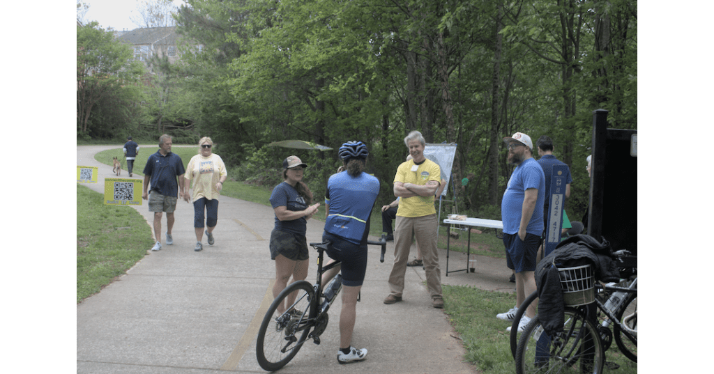 A group of people, one on a bicycle, stand around a table set up on the Silver Comet Trail