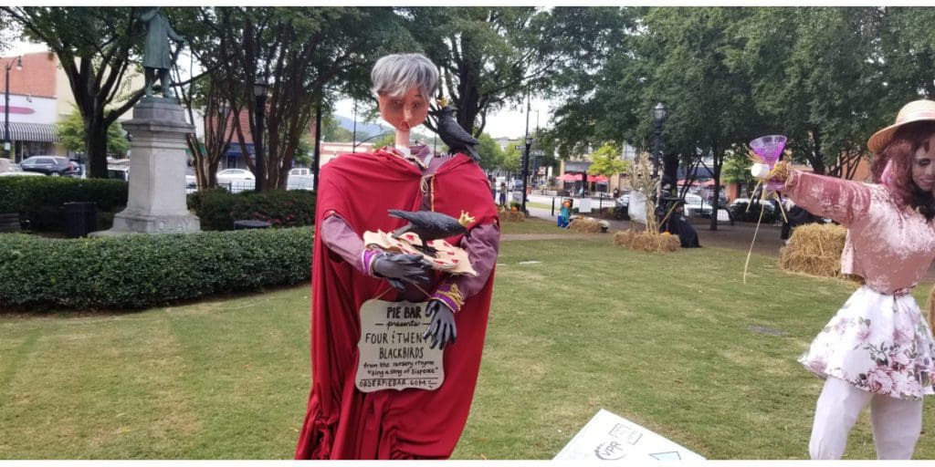 An elaborate scarecrow with a mannequin head holding a blackbird and a sign with a few words from the nursery rhyme (four and twenty blackbirds)