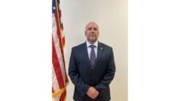 GBI Inspector Trent Hillsman wearing a dark suit and tie, poses with hands crossed in front next to an American flag